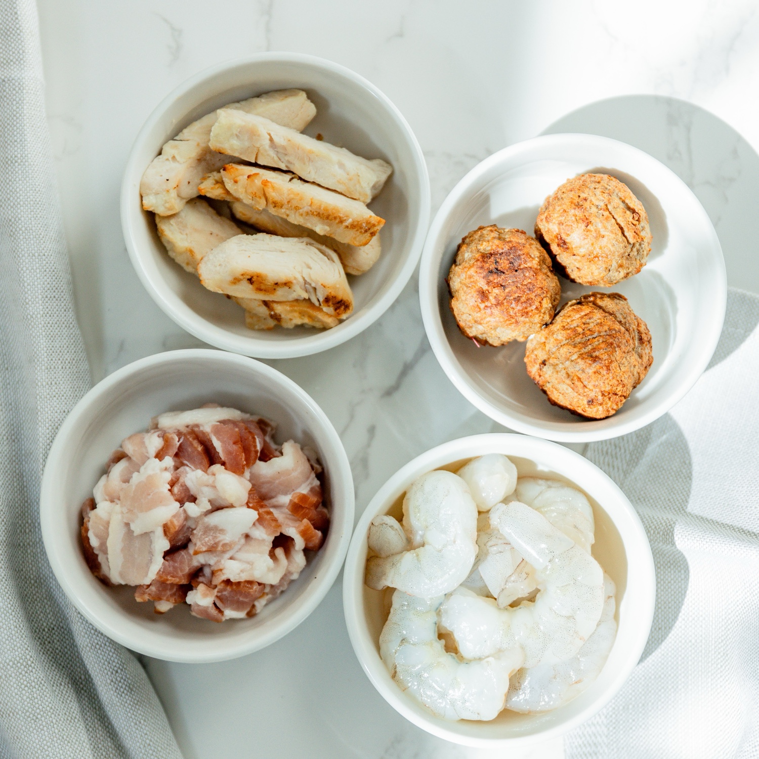 Bowls of grilled chicken, meatballs, bacon, and shrimp ready to add to a build-your-own pasta bowl